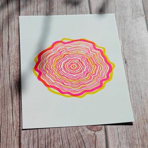 The A5 riso print at an angle on weathered wood in soft sunlight, concentric tree rings in pink and yellow