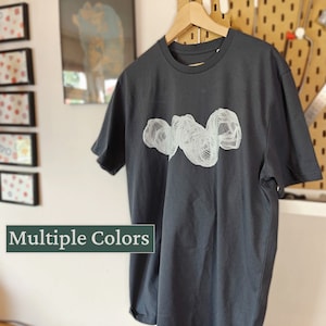 Könnte beinhalten: Ein dunkelgraues T-Shirt mit einem weißen grafischen Design aus einer Reihe abstrakter Formen. Das Design ist mittig auf der Vorderseite des Shirts platziert.