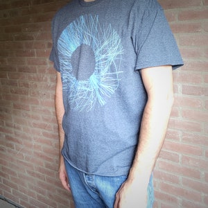 Side view of a man wearing the dark grey t-shirt with the blue radial line ring print against a brick wall