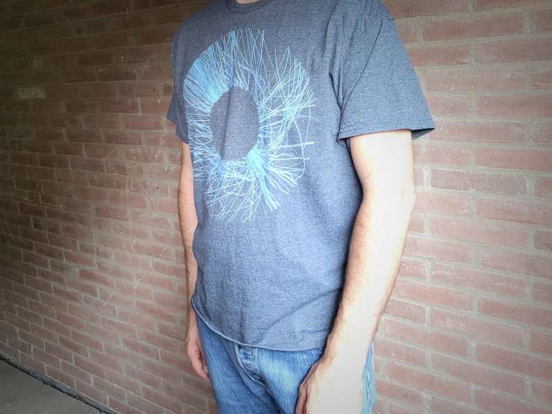Side view of a man wearing the dark grey t-shirt with the blue radial line ring print against a brick wall