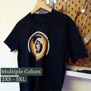 Può includere: Una t-shirt blu scuro con un design circolare multicolore. Il design presenta cerchi concentrici in tonalità di arancione, giallo e marrone. La t-shirt è disponibile nelle taglie 2XS-5XL. Colori multipli.