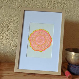 The A5 riso print in a light wooden frame on a shelf next to a singing bowl, concentric tree rings in pink and yellow
