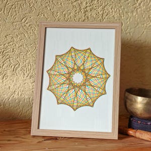 May include: A framed artwork with a geometric design in a light wood frame. The artwork showcases a star-shaped pattern with intersecting lines in yellow, orange, blue, and green. The artwork is displayed on a wooden surface.