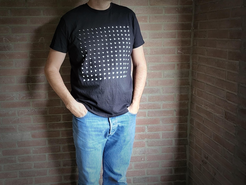 Man wearing a black organic cotton t-shirt with a grid of small white triangles, standing against a brick wall