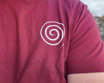 May include: A maroon short-sleeved t-shirt featuring a white graphic of a spiral on the left chest area. The spiral design has a textured, hand-drawn appearance with a small tail extending from the bottom right of the circular pattern.