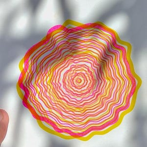 Three-panel photo collage of the A5 riso tree rings print — held outdoors top, close-up of the pink, yellow and orange concentric ring shapes in sunlight middle, framed on a shelf next to a singing bowl bottom
