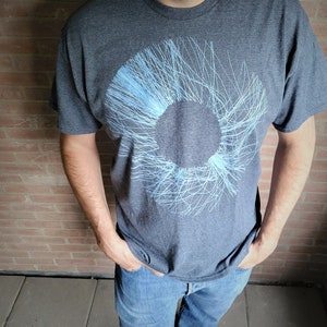 Man in a dark grey t-shirt with the blue radial line print, hands in his pockets against a brick wall