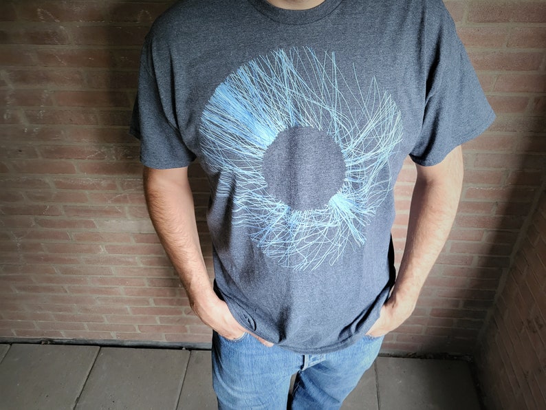 Man in a dark grey t-shirt with the blue radial line print, hands in his pockets against a brick wall