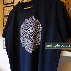 Op de afbeelding: Een donkerblauw t-shirt met een wit geometrisch ontwerp van concentrische cirkels. Het ontwerp is gecentreerd op het shirt en heeft een spiraaleffect. De tekst "multiple colors" is zichtbaar in de rechterbenedenhoek van de afbeelding.