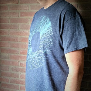 Man wearing a dark grey t-shirt in front of a brick wall; the print shows a wide ring of blue scribbled lines radiating from the centre, a procedurally generated pattern