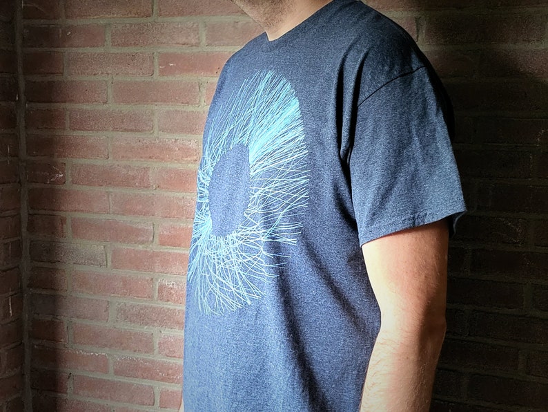 Man wearing a dark grey t-shirt in front of a brick wall; the print shows a wide ring of blue scribbled lines radiating from the centre, a procedurally generated pattern