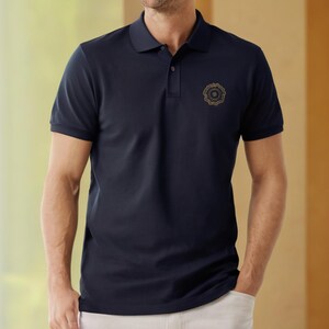 Op de afbeelding: Een donkerblauw poloshirt met een geborduurd logo in goud op de linkerborst. Het logo is een cirkelvormig ontwerp met ingewikkelde details.