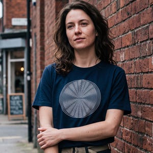 May include: A dark navy blue short-sleeved t-shirt featuring a large, centered white graphic print of a circular moiré pattern. The design consists of fine, concentric lines that create an optical illusion of depth and movement on the chest.