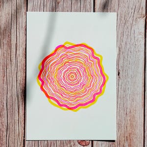 The full A5 riso print flat on wood in dappled shade, concentric tree rings in pink and yellow