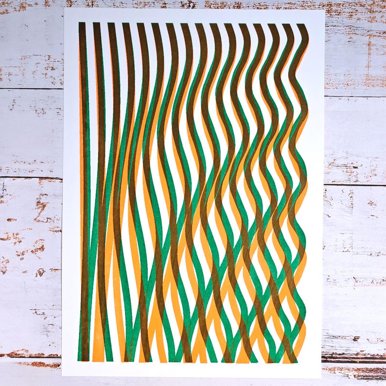 Handmade A3 risograph print with wavy green, yellow-orange and brown lines lying flat on a whitewashed wooden surface