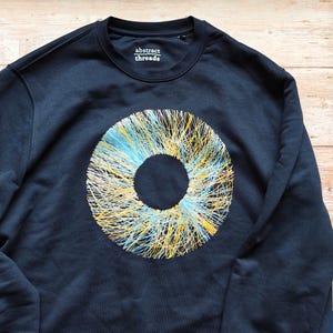 Op de afbeelding: Marineblauw sweatshirt met een groot cirkelvormig ontwerp in het midden, bestaande uit gele, blauwe en witte lijnen. De tekst "abstract threads" is op de borst gedrukt.