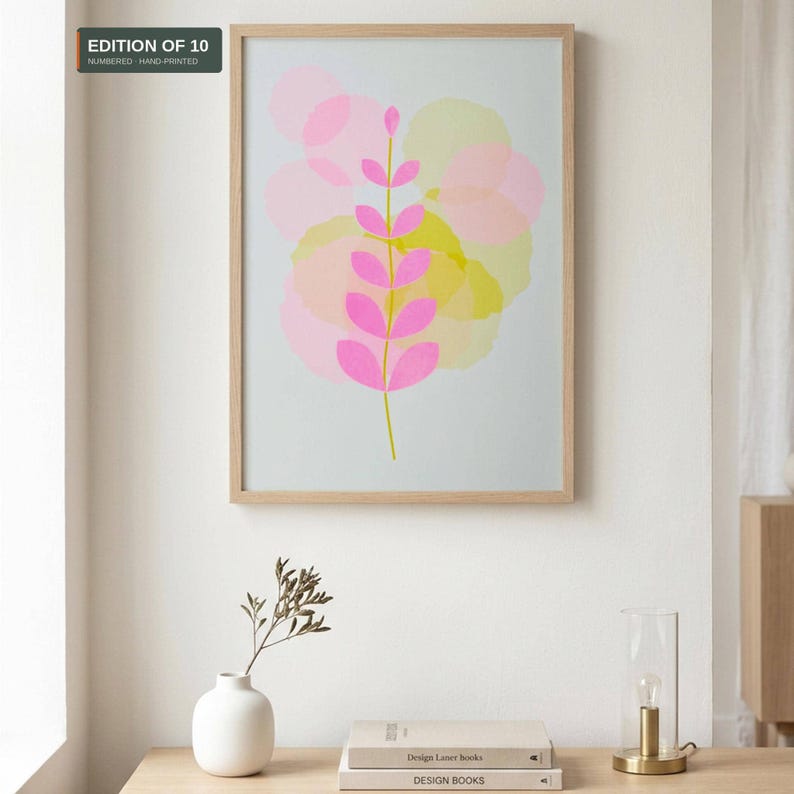 Framed A3 botanical riso print with a pink leaf sprig over pastel circles, in an oak frame on a white wall above a sideboard with books and a vase