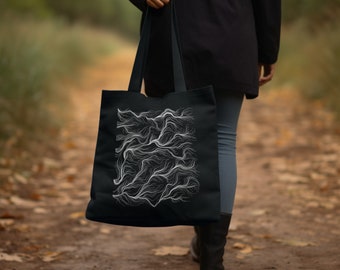 Può includere: Una borsa tote nera con un designo di arte astratta a linee bianche. La borsa viene portata da una persona che cammina su un sentiero.