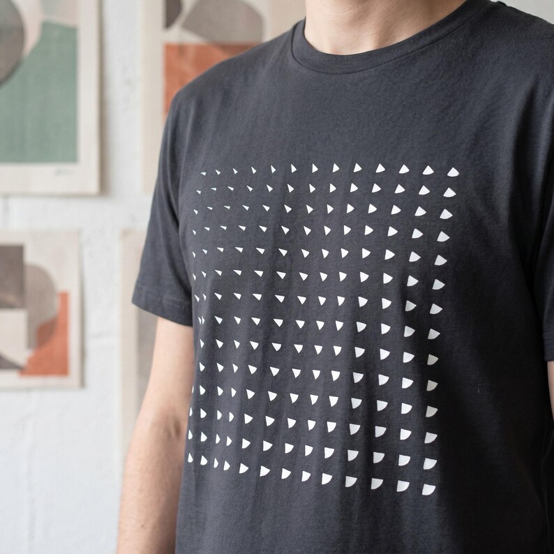 Man wearing the india ink grey t-shirt with the white rotating triangle pattern, white wall with art prints behind