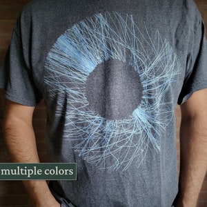 Puede incluir: Una camiseta gris oscuro con un diseño circular blanco y azul claro. El diseño está formado por muchas líneas finas que irradian hacia afuera desde el centro.
