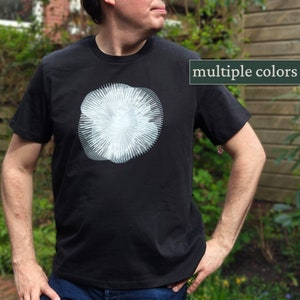 Könnte beinhalten: Ein schwarzes T-Shirt mit einer weißen Grafik eines stilisierten Pilzes. Der Text "multiple colors" befindet sich in einem grünen Kasten in der oberen rechten Ecke des Bildes.