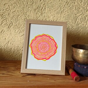 Framed A5 riso print standing on a wooden shelf against a textured wall, concentric tree rings in pink and yellow