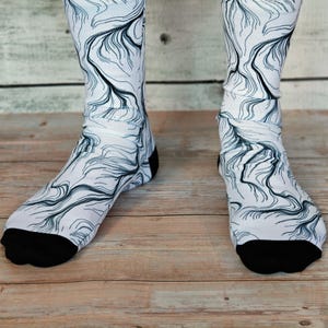 Peut inclure: Paire de chaussettes blanches avec un motif abstrait noir et gris. Les talons et les orteils sont noirs. Les chaussettes sont posées sur une surface en bois.