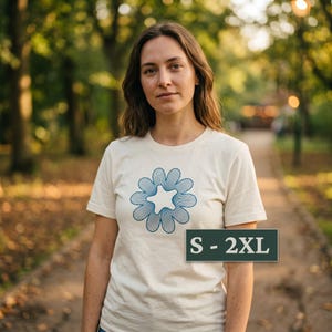May include: A cream-colored short-sleeve t-shirt featuring a blue graphic of a stylized flower with a star-shaped center. A dark green rectangular label is overlaid on the lower right side with the white text S - 2XL indicating size range.