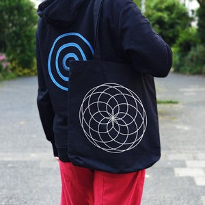 May include: A black tote bag with a white geometric design. The bag has a long strap and is being carried by a person wearing a black hoodie with a blue circle design.