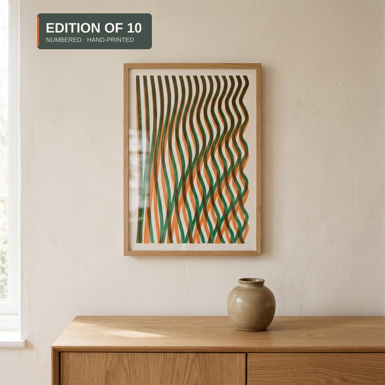 Framed A3 risograph print with wavy green, orange and brown lines in a thin oak frame on a warm white wall above an oak sideboard with a ceramic vase.
