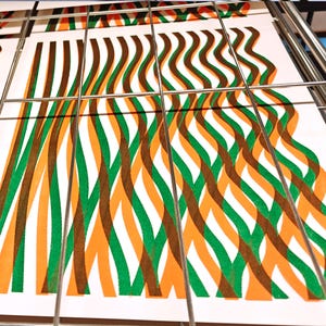 Freshly printed A3 risograph prints with green and orange wavy lines drying on a metal rack in the print studio