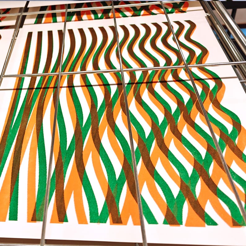Freshly printed A3 risograph prints with green and orange wavy lines drying on a metal rack in the print studio