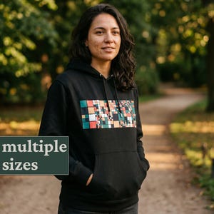 Puede incluir: Sudadera con capucha negra con un diseño geométrico colorido en el pecho. El diseño presenta cuadrados en tonos turquesa, naranja y crema. La sudadera tiene un bolsillo delantero y una capucha. El texto "multiple sizes" está visible.