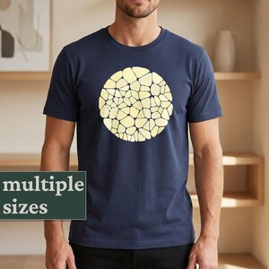 Puede incluir: Camiseta azul marino con un diseño abstracto circular grande en tonos amarillos. El diseño presenta un patrón de formas irregulares. Las palabras "multiple sizes" se muestran en un rectángulo verde.