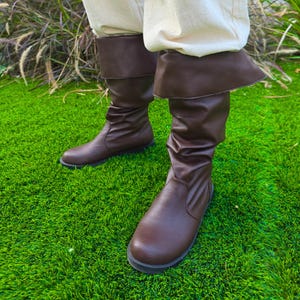 Puede incluir: Botas altas de cuero marrón con un puño doblado. Las botas tienen una textura ligeramente arrugada y una suela plana. Las botas se usan sobre pantalones de color claro y están sobre hierba verde.