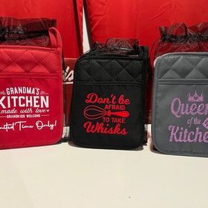 May include: Three quilted pot holders with different sayings. The red pot holder says "Grandma's Kitchen made with love - Grandkids Welcome - Limited Time Only!" The black pot holder says "Don't be afraid to take whisks". The gray pot holder says "Queen of the Kitchen" with a crown and a spatula.