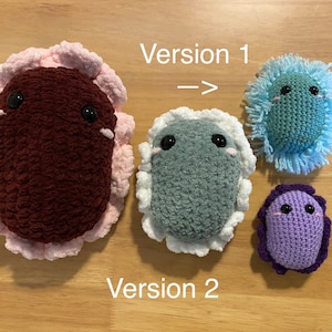May include: Three crocheted plush toys in different colors and sizes. The largest is maroon with a pink border, the middle is gray with a white border, and the smallest is purple with a dark purple border. The text "Version 1" and "Version 2" are visible in the image.
