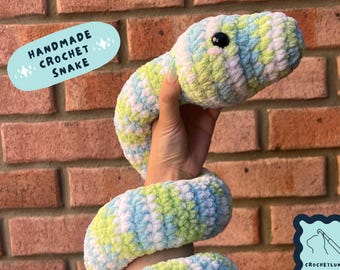 Peut inclure: Serpent en crochet fait main en vert, blanc et bleu. Le serpent est enroulé et a un œil noir. Le texte "HANDMADE CROCHET SNAKE" est dans un cercle bleu avec des étoiles blanches.