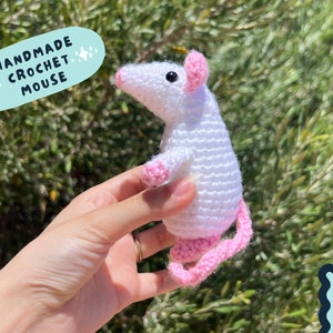 May include: Handmade crochet mouse with a white body and pink ears, nose, and tail. The mouse is being held in a hand against a green background. The text "HANDMADE CROCHET MOUSE" is in a blue speech bubble with white stars.