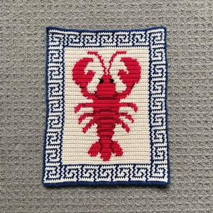 May include: A rectangular, handmade crochet piece featuring a red lobster design on a cream background. The lobster is centered within a blue and white Greek key border. The piece is likely a decorative item or a small mat.
