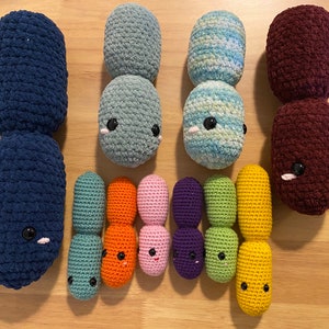 May include: A set of nine crocheted plush toys shaped like bacteria. The toys are in various colors, including blue, green, orange, pink, purple, and yellow. The toys have embroidered eyes and mouths.
