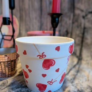 May include: A white ceramic plant pot adorned with red hearts and bows. The pot has a smooth finish and a simple, rounded form. The hearts are of varying sizes and are scattered across the pot's exterior. A charming, handmade item for home decor.