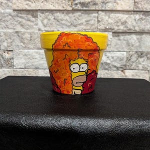 Puede incluir: Maceta amarilla pintada a mano con un personaje de dibujos animados con pelo naranja y expresión de sorpresa. La maceta tiene una base amarilla con detalles naranjas y rojos, perfecta para añadir un toque de diversión a cualquier espacio.