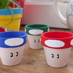 Puede incluir: Tres pequeñas macetas con forma de hongo pintadas a mano. Cada maceta tiene un borde de diferente color: azul, verde y rojo, con lunares blancos. Las macetas tienen una base rosa claro con dos detalles negros en forma de ojo.