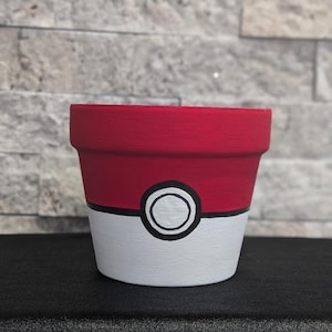 Op de afbeelding: Handbeschilderde keramische bloempot in de stijl van een Poké Ball. De bovenkant is rood, de onderkant is wit en in het midden bevindt zich een zwart-wit cirkelontwerp. De pot is bedoeld voor het planten van bloemen of andere planten.