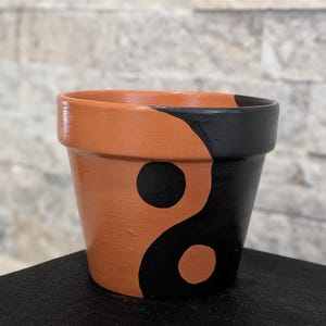 Può includere: Un vaso di fiori in ceramica color terracotta con un design yin-yang. Il vaso è diviso verticalmente, con una metà dipinta di nero e l'altra di terracotta. Il simbolo yin-yang è formato da due cerchi neri sul lato terracotta.