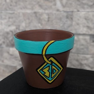 Puede incluir: Una maceta de cerámica marrón con un borde turquesa y un gráfico amarillo y negro. El gráfico tiene forma de diamante con las letras "3D" en su interior. La maceta está sobre una superficie negra.
