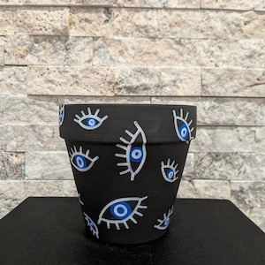 Puede incluir: Maceta de cerámica negra decorada con diseños de ojos blancos y azules. La maceta es cilíndrica y está sobre una superficie negra, con una pared de piedra texturizada de fondo.