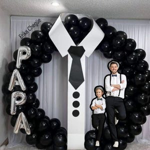 May include: A balloon arch in the shape of a shirt and tie, with the word "PAPA" in silver balloons. The arch is surrounded by black balloons. A man and a child stand in front of the arch, dressed in formal attire.