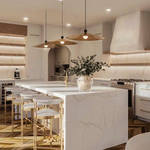 May include: A kitchen island with a white marble countertop and base, featuring three gold-toned pendant lights above. Four white and gold bar stools are arranged along the side. A clear glass vase with green foliage sits on the island surface.